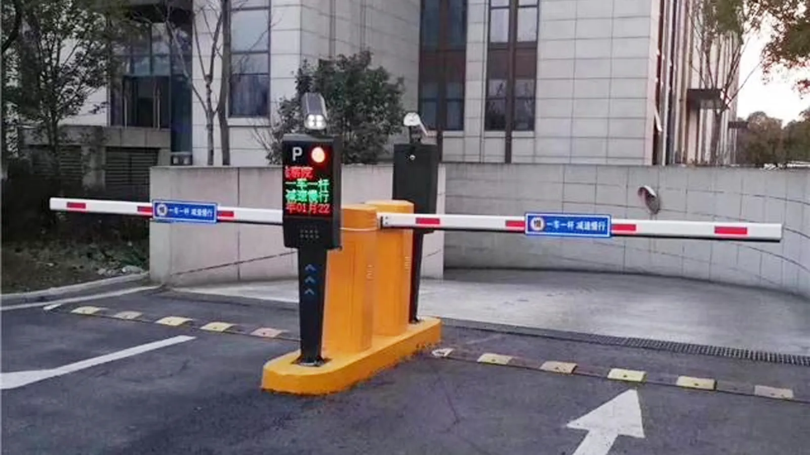 Automatic parking barrier gate system at a building entrance, featuring dual boom barriers, a control pedestal with LED display, and lane markings.
